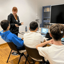 Laboratorio di storie multimediali nel nodo Galattica di Trani tre ragazzi seduti di spalle con una ragazza che parla con loro in un stanza con scrivania, monitor e scaffali.