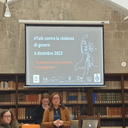 Foto dall'evento sulla sensibilizzazione contro la violenza di genere Donna che parla in piedi, una sadura al lato e una dietro in sala con scaffali e telo su cui vengono proiettate slide