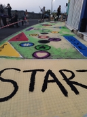 dalla parte destra dell'immagine c'è un marciapiede che è stato dipinto. All'inizio c'è una scritta "START", a destra una scritta "love" verticale su uno sfondo blu. Nel centro vari simboli di varie forme e colori. Dall'altro della strada alcuni ragazzi seduti e in piedi