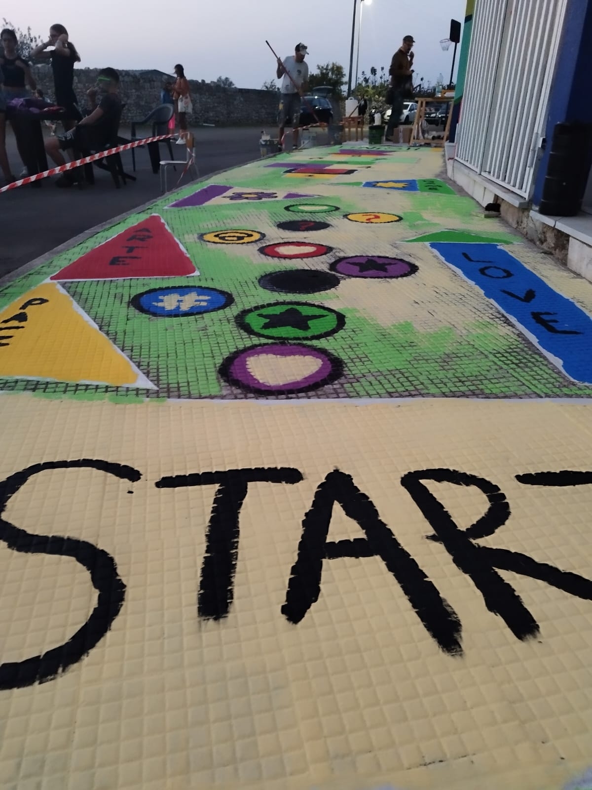 dalla parte destra dell'immagine c'è un marciapiede che è stato dipinto. All'inizio c'è una scritta "START", a destra una scritta "love" verticale su uno sfondo blu. Nel centro vari simboli di varie forme e colori. Dall'altro della strada alcuni ragazzi seduti e in piedi