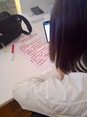 Laboratorio del Piano Regionale "Think Europe" al nodo San Pancrazio Salentino Una ragazza con capelli castani e maglia bianca, vista di spalle, usa un cellulare mentre su un tavolo bianco davanti a lei c'è un foglio con scritte a mano di colore rosso, il pennarello rosso, una borsetta nera e in fondo una calcolatrice.