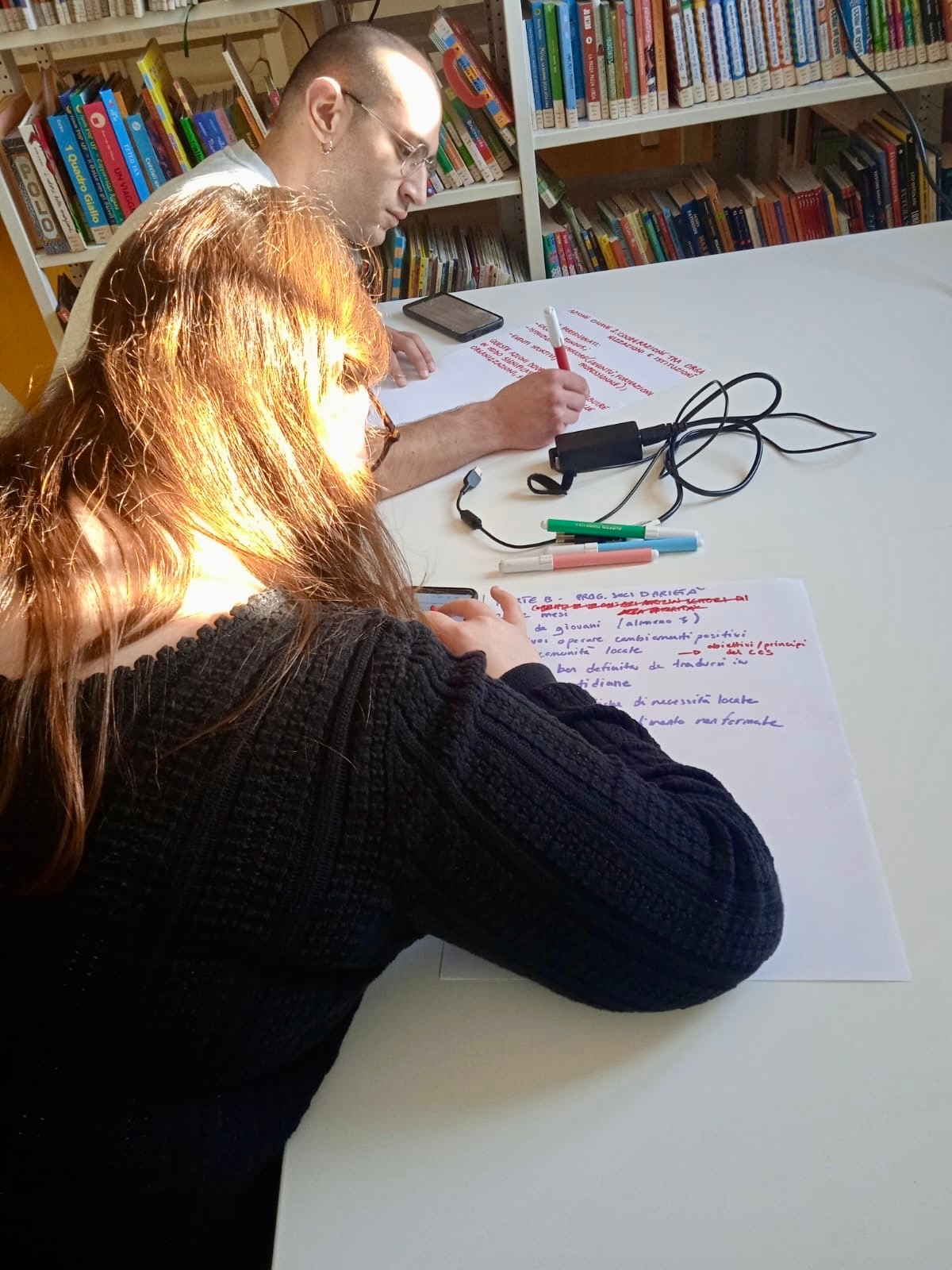 Una ragazza con gli occhiali, vista di spalle, scrive su un foglio su un tavolo bianco e accanto a lei un ragazzo con gli occhiali scrive su un altro foglio con un pennarello rosso, in mezzo a loro un alimentare nero da pc col cavo e altri tre pennarelli, in fondo una libreria piena di volumi.