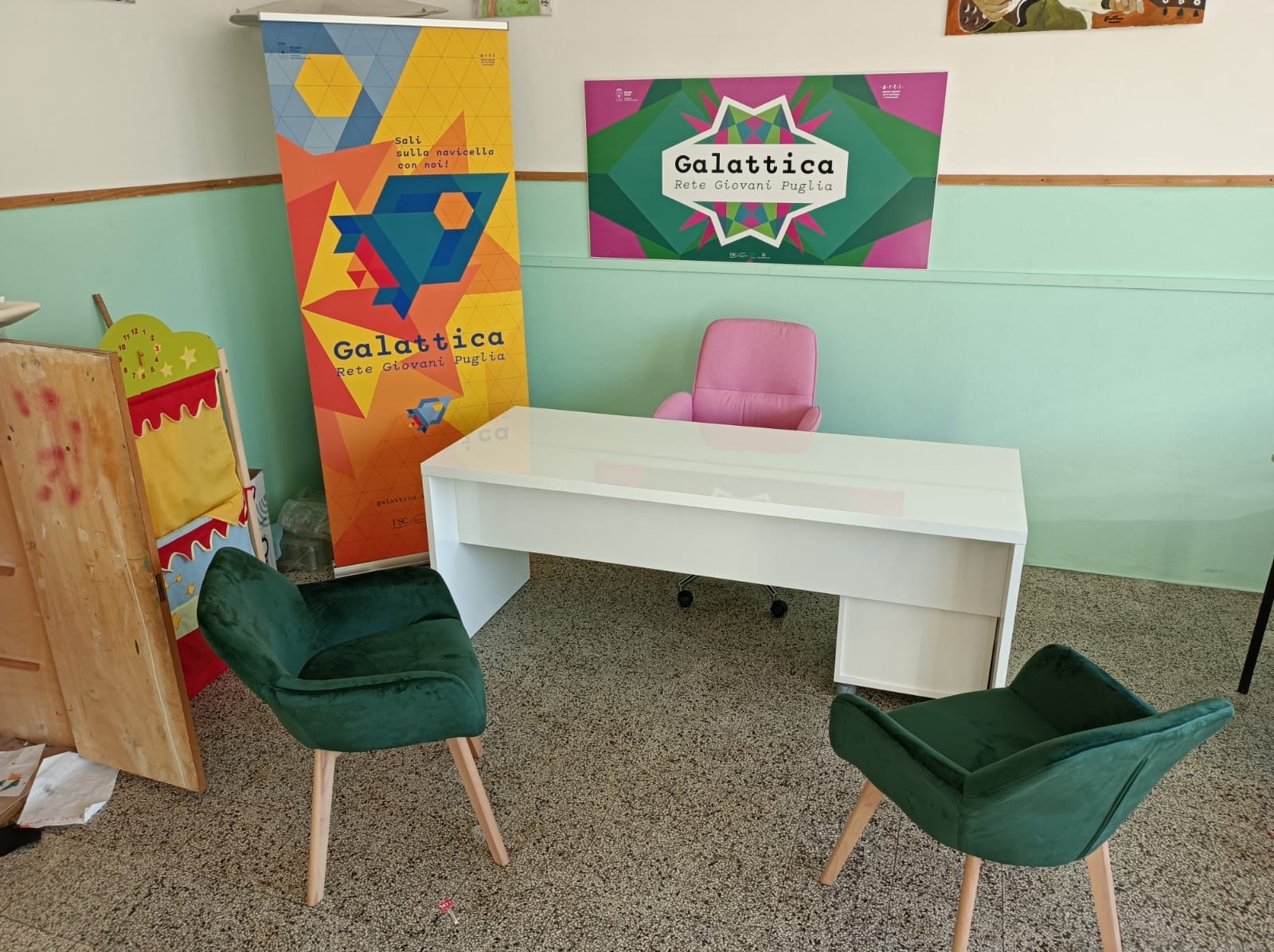 Una stanza con una scrivania, sedie e un poster verde con la scritta "Galattica"