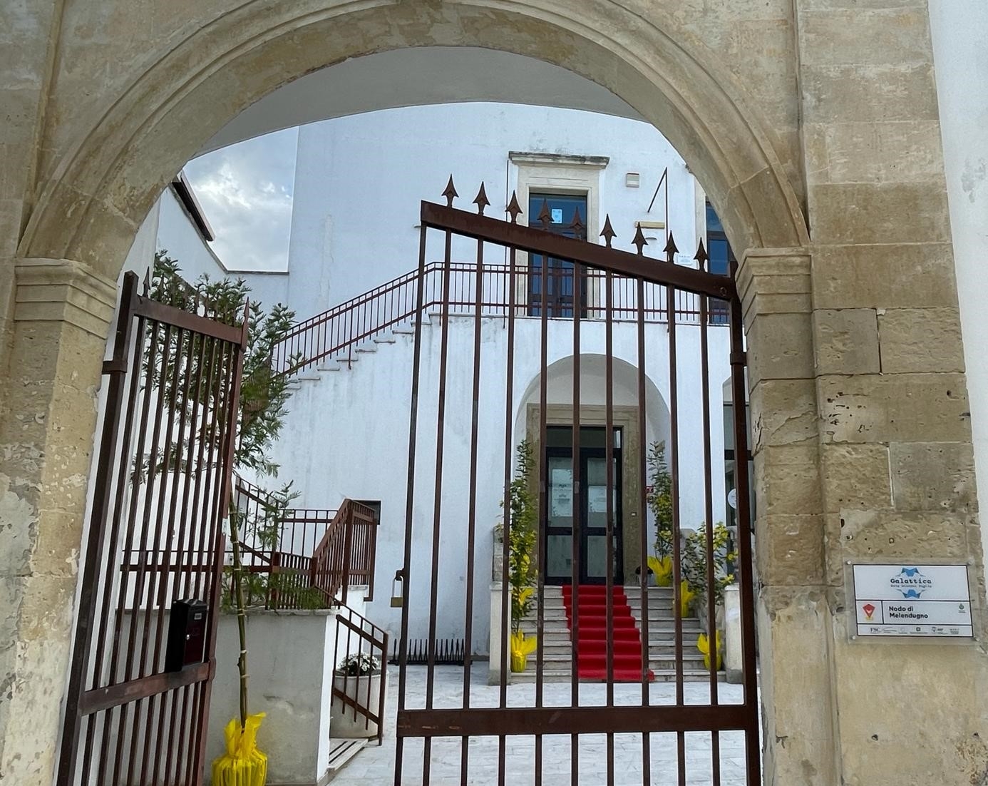 Cancello aperto in un arco in pietra, di cui in una colonna a destra c'è una targa del nodo di Galattica di Melendugno, e in fondo il cortile di un palazzo storico dipinto di bianco con una scalinata che arriva alla porta del piano rialzato e un'altra scala laterale alla porta del primo piano.