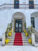 Entrata della sede del nodo di Melendugno all'inaugurazione di Galattica. Scalinata con tappeto rosso al centro e ai lati vasi gialli con alberelli, la quale arriva a una porta a vetro sotto un arco nell'edificio dipinto di bianco, e in alto si vedono una inferriata e dietro un'altra porta al primo piano.