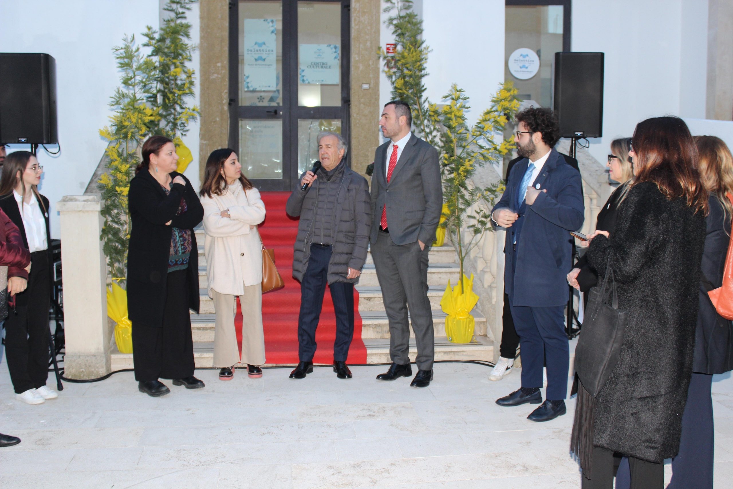 Persone in piedi davanti alla scalinata con un tappeto rosso e degli alberelli, la quale arriva alla porta a vetri del piano rialzato.