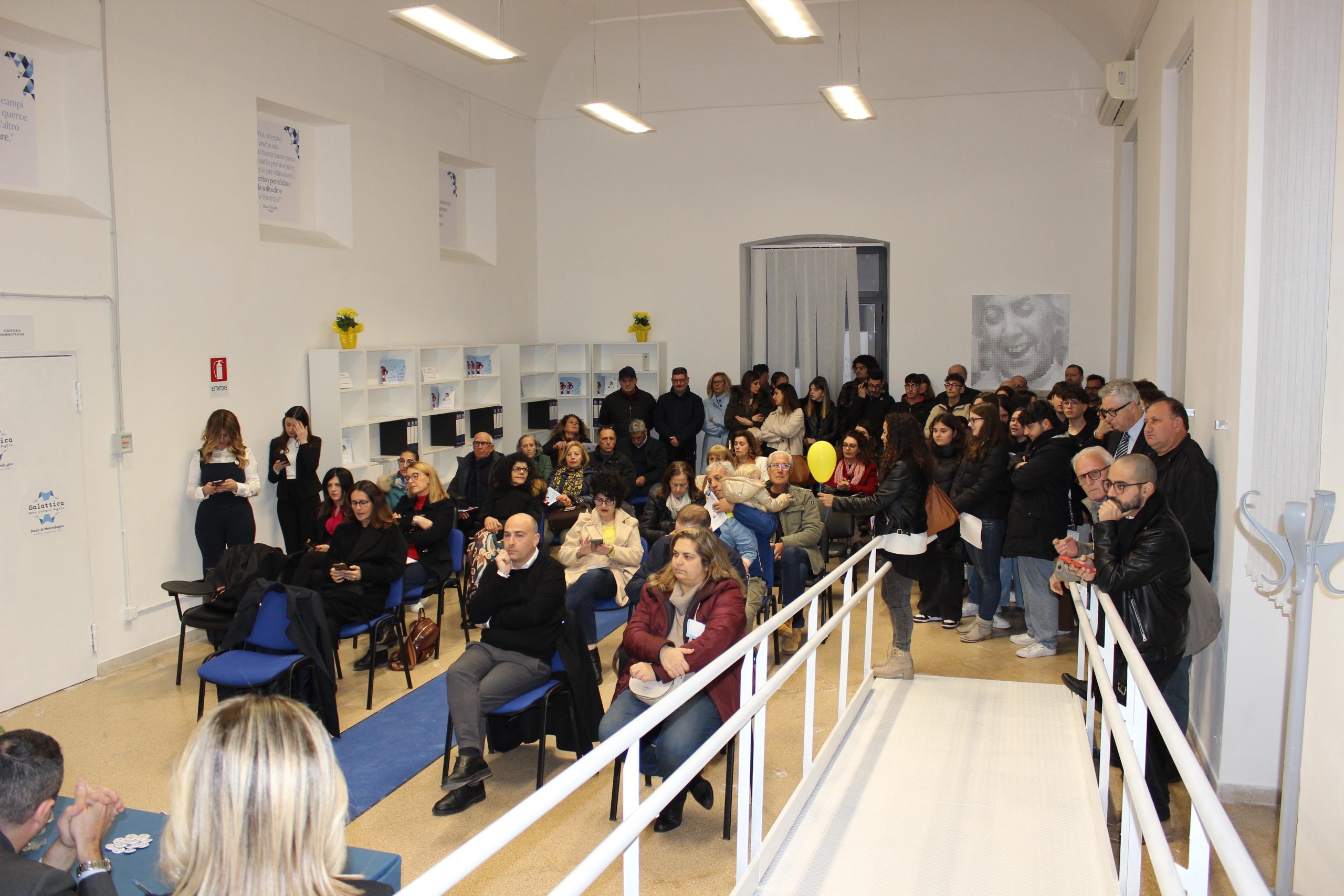 Persone sedute in platea in un'ampia sala dalle mura bianche e in primo piano a destra una rampa bianca con corrimano.