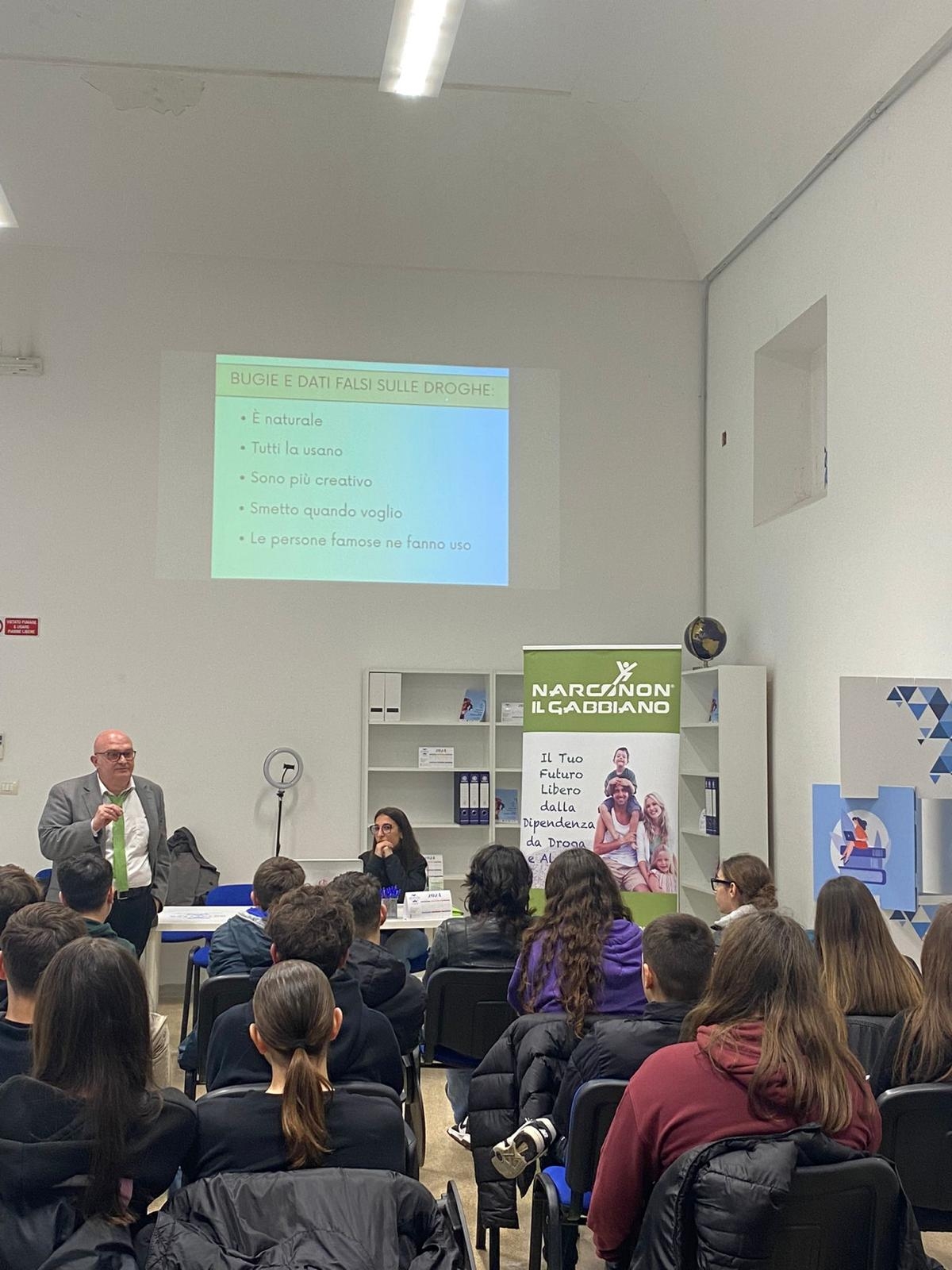 In una sala giovani seduti, visti di spalle, ascoltano un uomo in piedi che parla e sullo sfondo in alto viene proiettata una slide sul muro bianco.