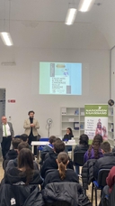 Incontro "Prevenzione e futuro: falsi miti e credenze sulle droghe" In una sala giovani seduti, visti di spalle, ascoltano un uomo in piedi che parla con un altro uomo che gli sta accanto in piedi e sullo sfondo in alto viene proiettata una slide sul muro bianco.