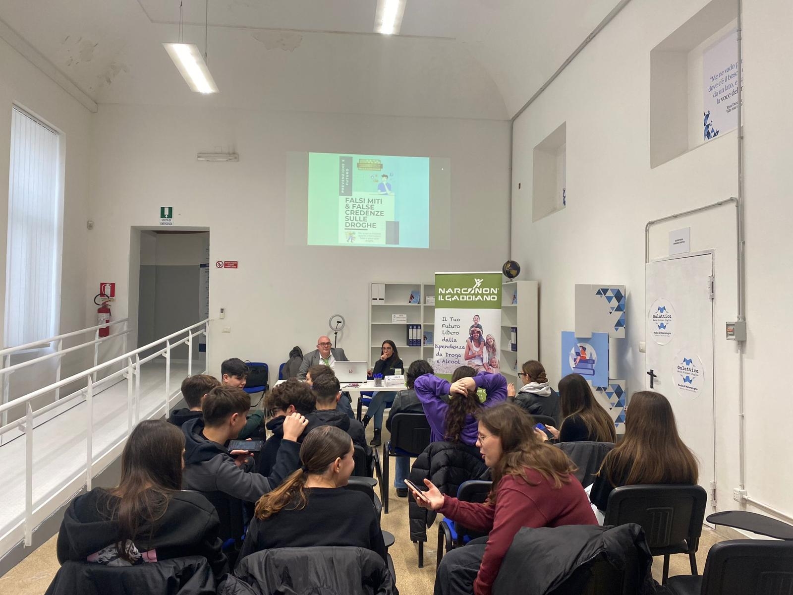 In una sala giovani seduti, visti di spalle, sono di fronte a due persone sedute dietro a un tavolo bianco sullo sfondo e dietro di loro in alto viene proiettata una slide sul muro bianco.