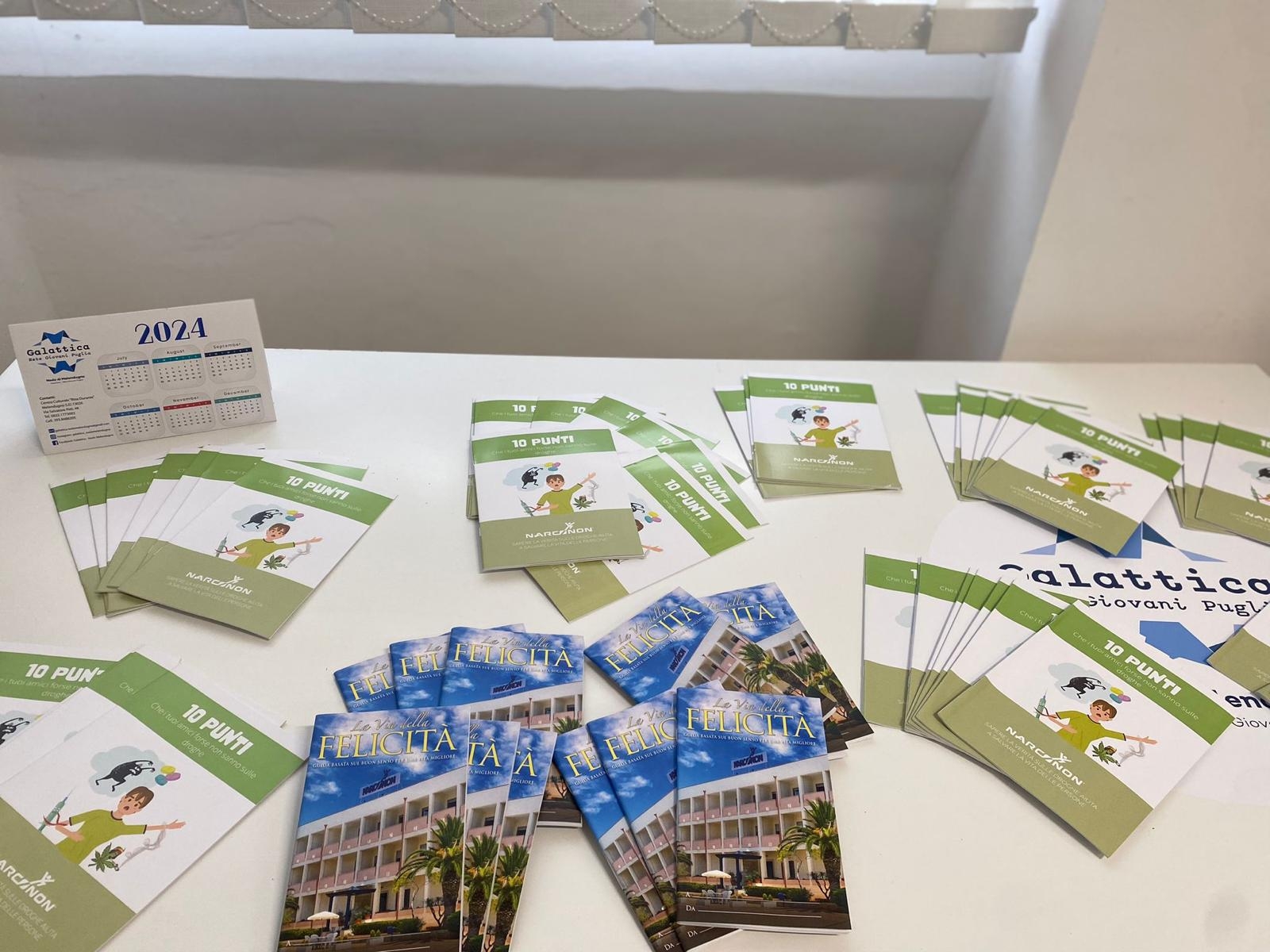 Due diversi tipi di brochure disposte su un tavolo bianco con un calendario da tavolo con il logo del nodo di Galattica di Melendugno.