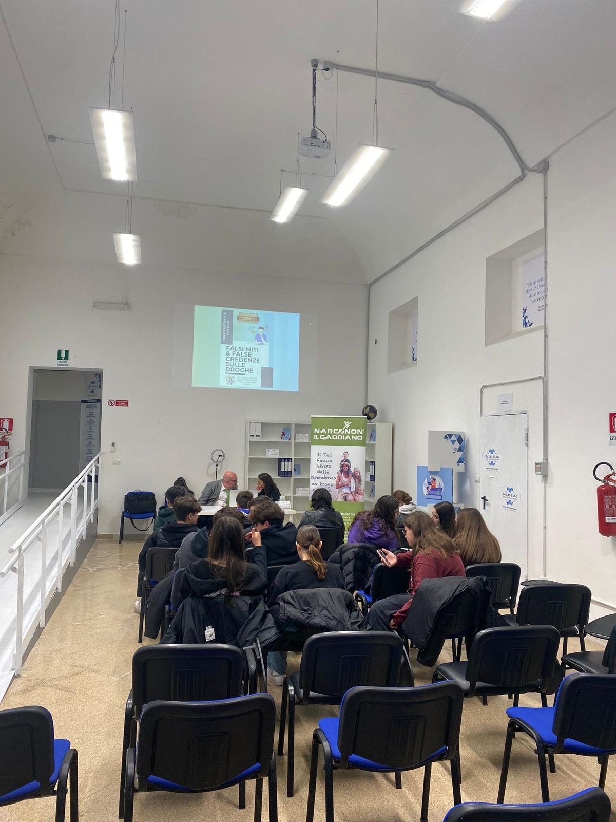 In una sala giovani seduti, visti di spalle, sono di fronte a due persone sedute dietro a un tavolo bianco sullo sfondo e dietro di loro in alto viene proiettata una slide sul muro bianco.