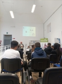 Incontro "La verità sulle droghe" Giovani seduti, visti di spalle, sono di fronte a un uomo in piedi che parla accanto a una ragazza seduta dietro a una scrivania con pc.