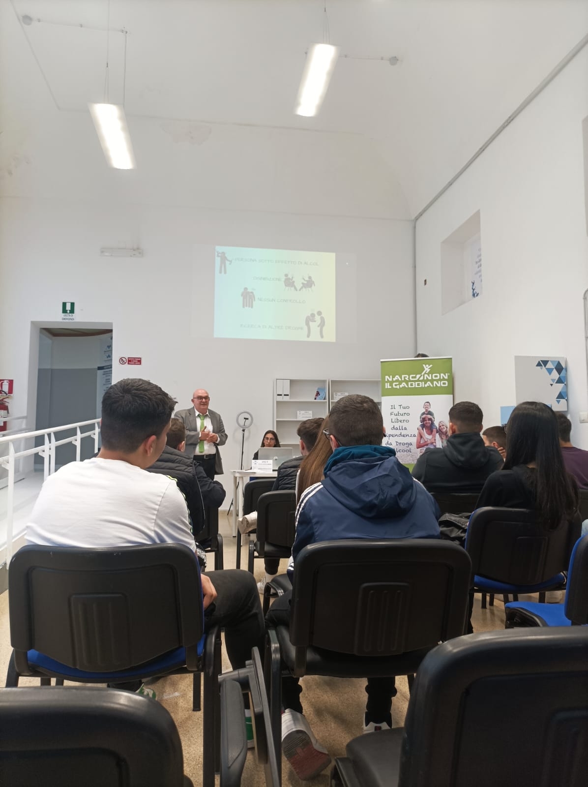 Giovani seduti, visti di spalle, sono di fronte a un uomo in piedi che parla accanto a una ragazza seduta dietro a una scrivania con pc.