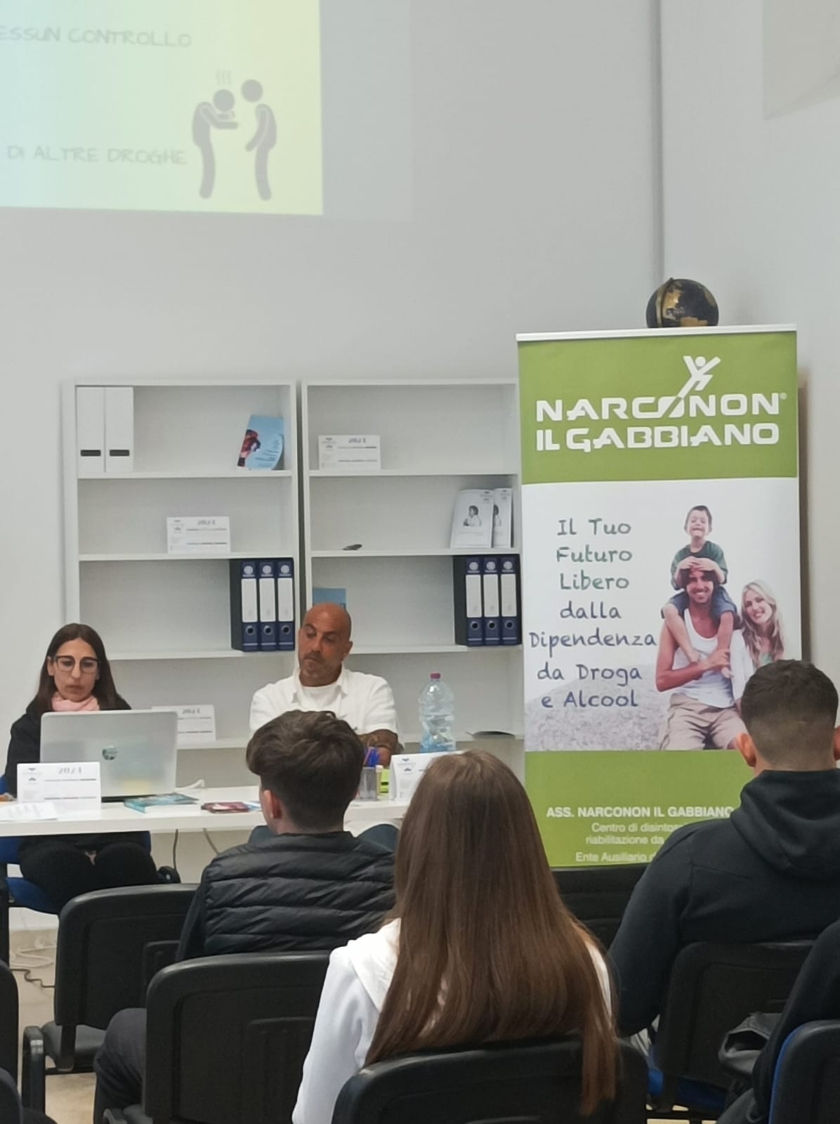 Giovani seduti, visti di spalle, che si trovano di fronte a due persone sedute dietro a un tavolo bianco con un pc e accanto un roll up.