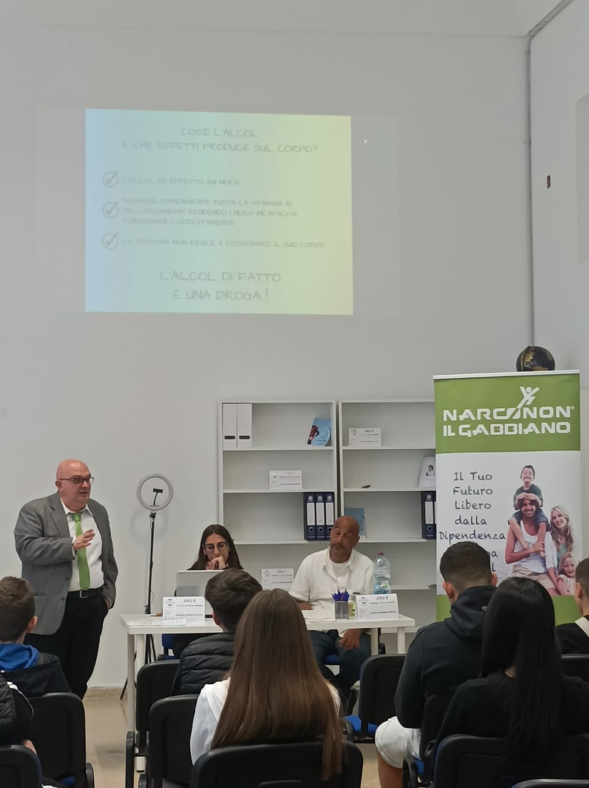 Giovani seduti, visti di spalle, si trovano di fronte a due persone sedute dietro a un tavolo bianco con un pc e a destra un roll up e a sinistra un uomo in piedi.