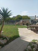 Spazi esterni del nodo di Melendugno Giardino con strada al centro, tavolo in legno sulla destra e in fondo un forno in pietra con cielo azzurro.