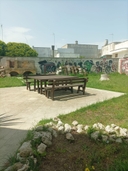 Spazi esterni del nodo di Melendugno Giardino esterno del nodo con tavolo e panche in legno, in fondo muro e forno in pietra con cielo azzurro.