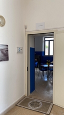 Entrata aula didattica del nodo di Melendugno Porta bianca aperta nel muro bianco, all'interno ci sono tavoli e sedie di un'aula didattica.