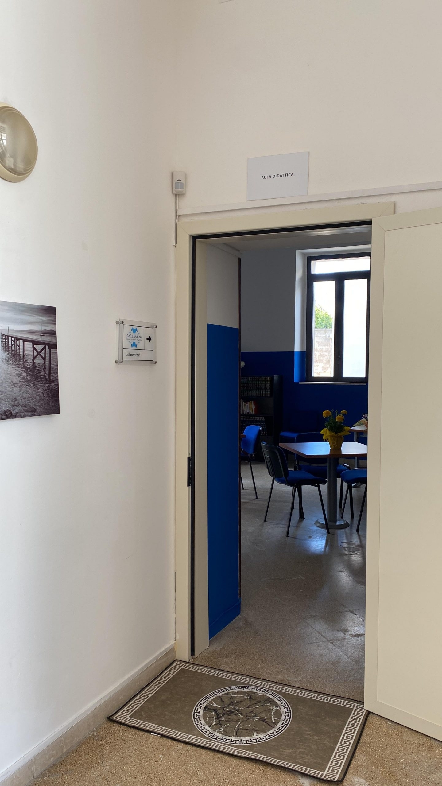 Porta bianca aperta nel muro bianco, all'interno ci sono tavoli e sedie di un'aula didattica.