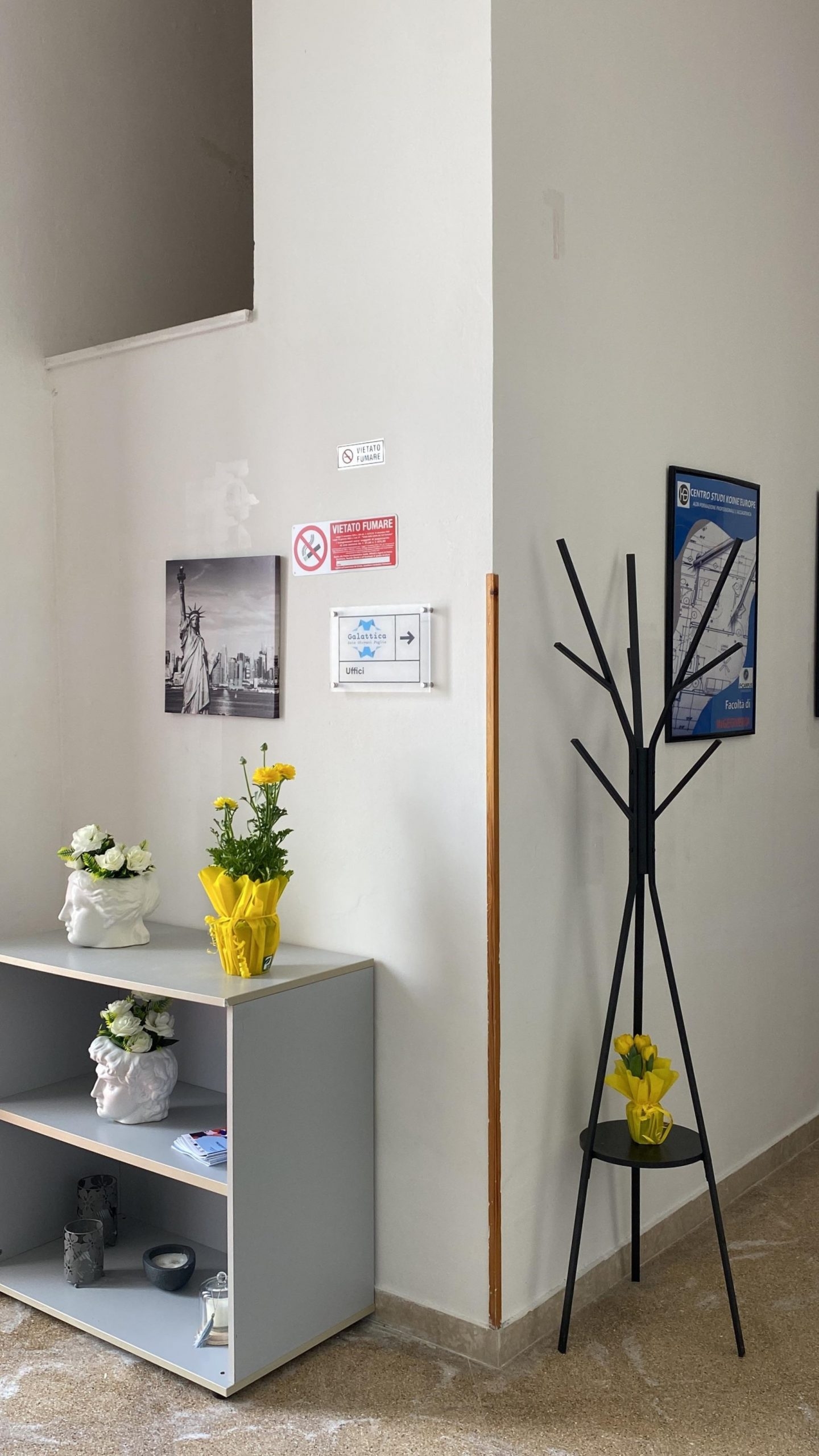 Angolo di un muro bianco all'interno dell'edificio, un mobiletto basso con vasi con fiori a sinistra e un appendiabiti a forma di albero a destra.