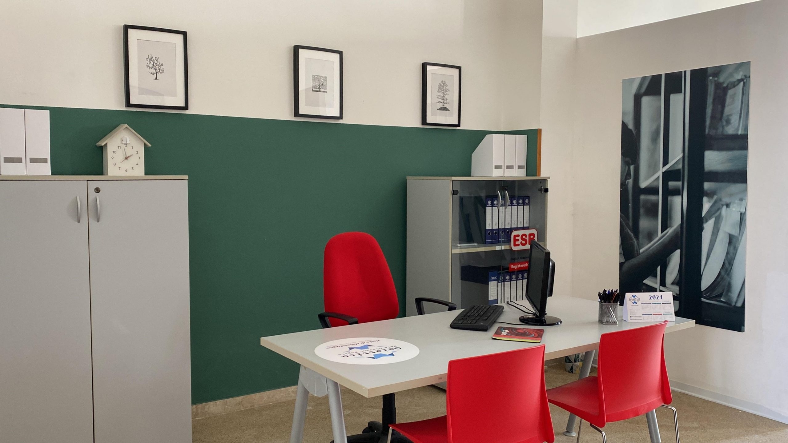 Ufficio con tavolo grigio e sedie rosse, un pc sul tavolo e a sinistra un mobiletto grigio, sullo sfondo il muro verde e bianco con tre piccoli quadri e un poster sulla destra.