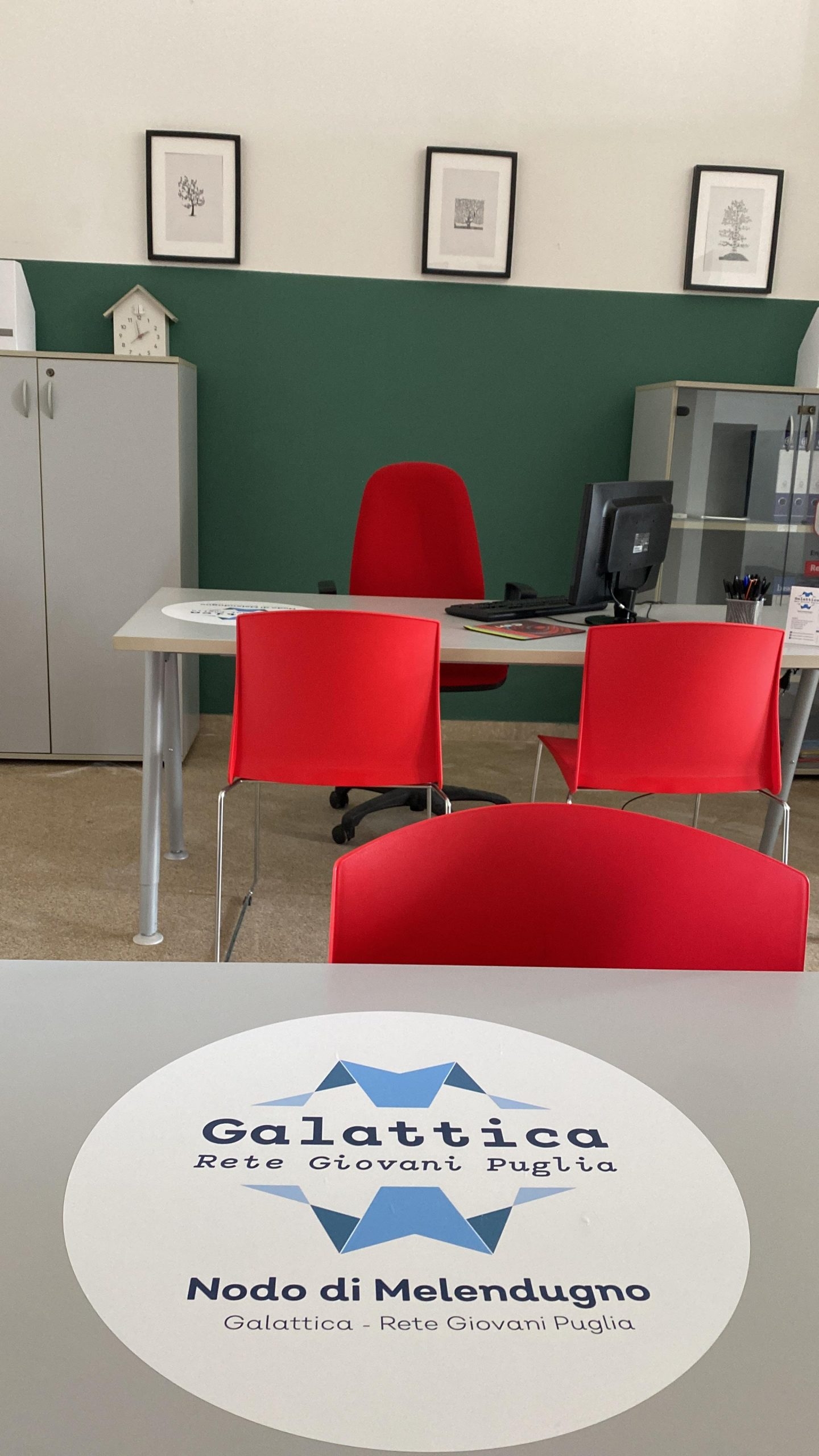 Tavolo grigio con logo del nodo di Melendugno e sedie rosse, sullo sfondo tavolo grigio, un armadietto grigio sulla sinistra e il muro verde e bianco con tre piccoli quadri.