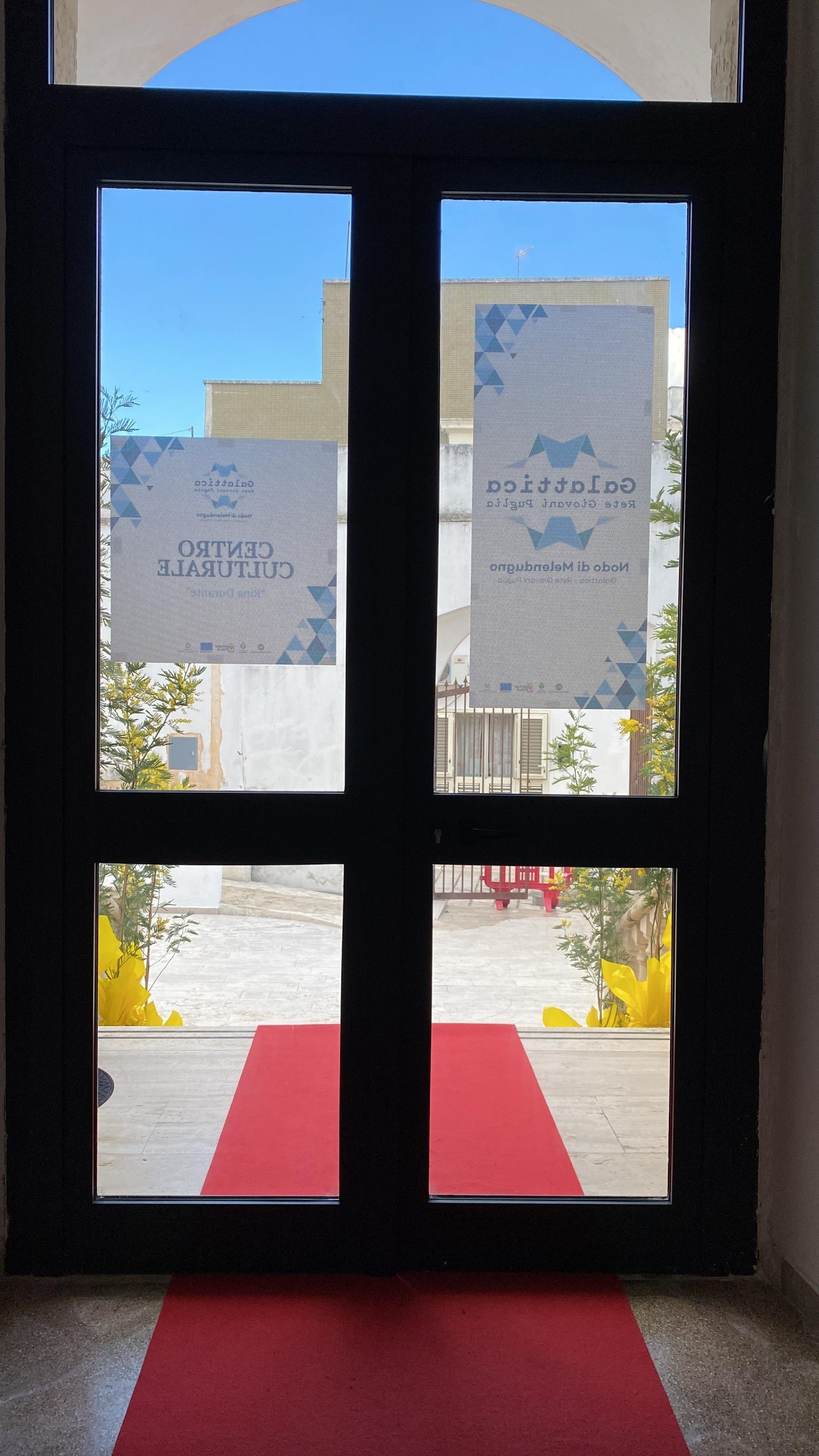 Vista interna di una porta a vetri con tappeto rosso che porta alle scale esterne del nodo di Melendugno.