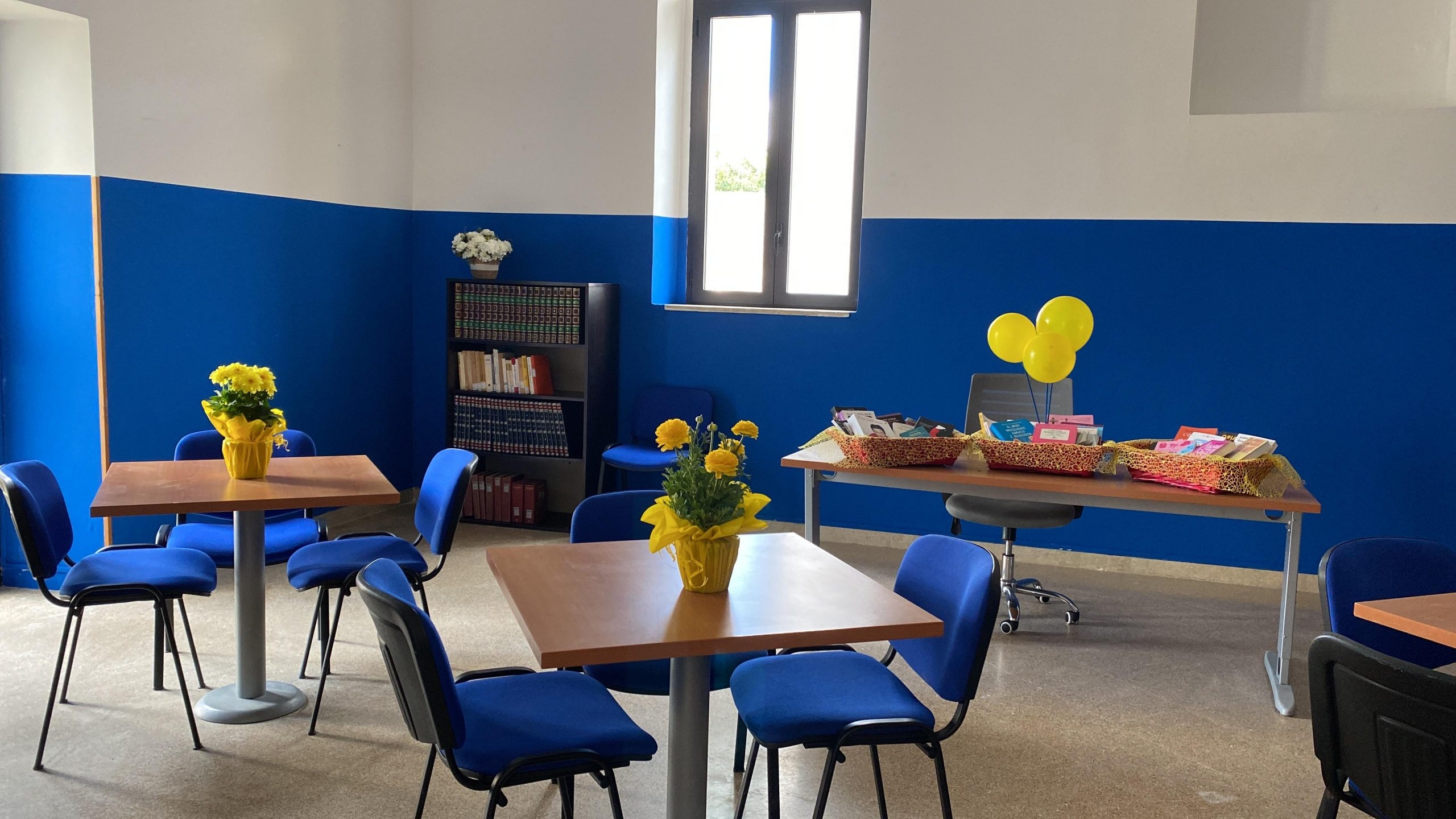 Aula studio con tavoli in legno, sedie blu e fiori gialli sui tavoli.