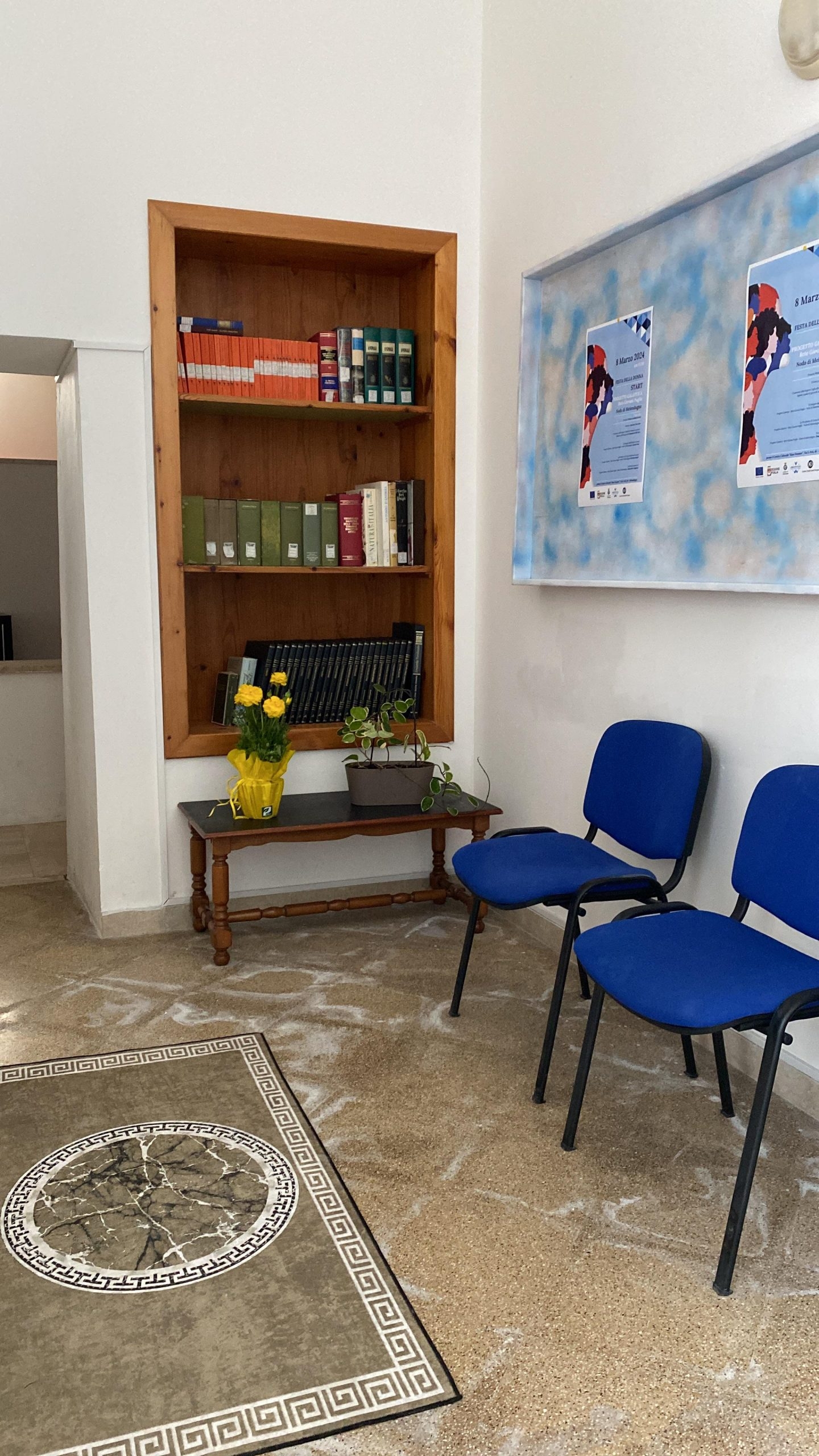 Sala con due sedie blu, due poster sul muro a destra e una libreria e un tavolo in legno di fronte.