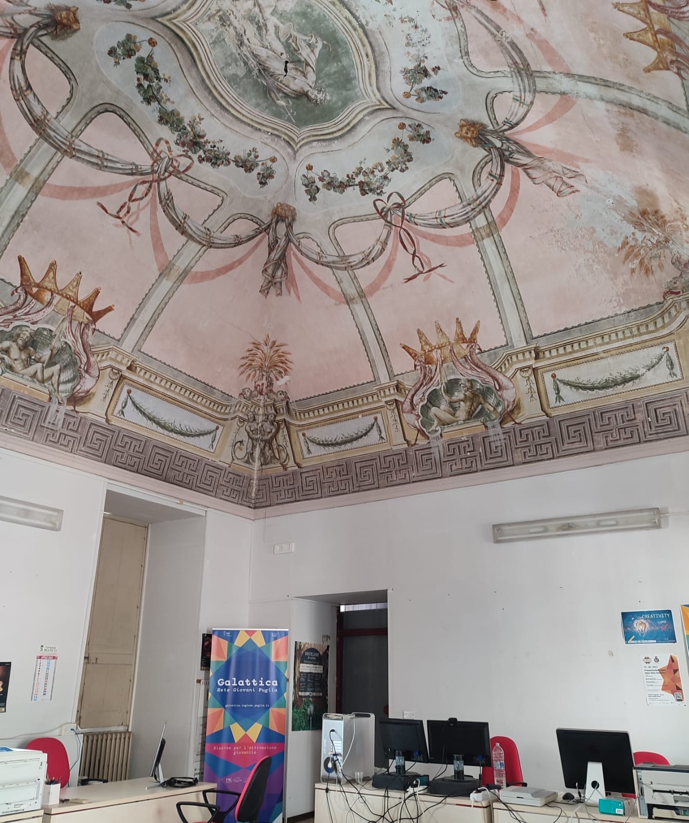sala con soffitto affrescato, tavoli con pc e roll up con il logo di Galattica
