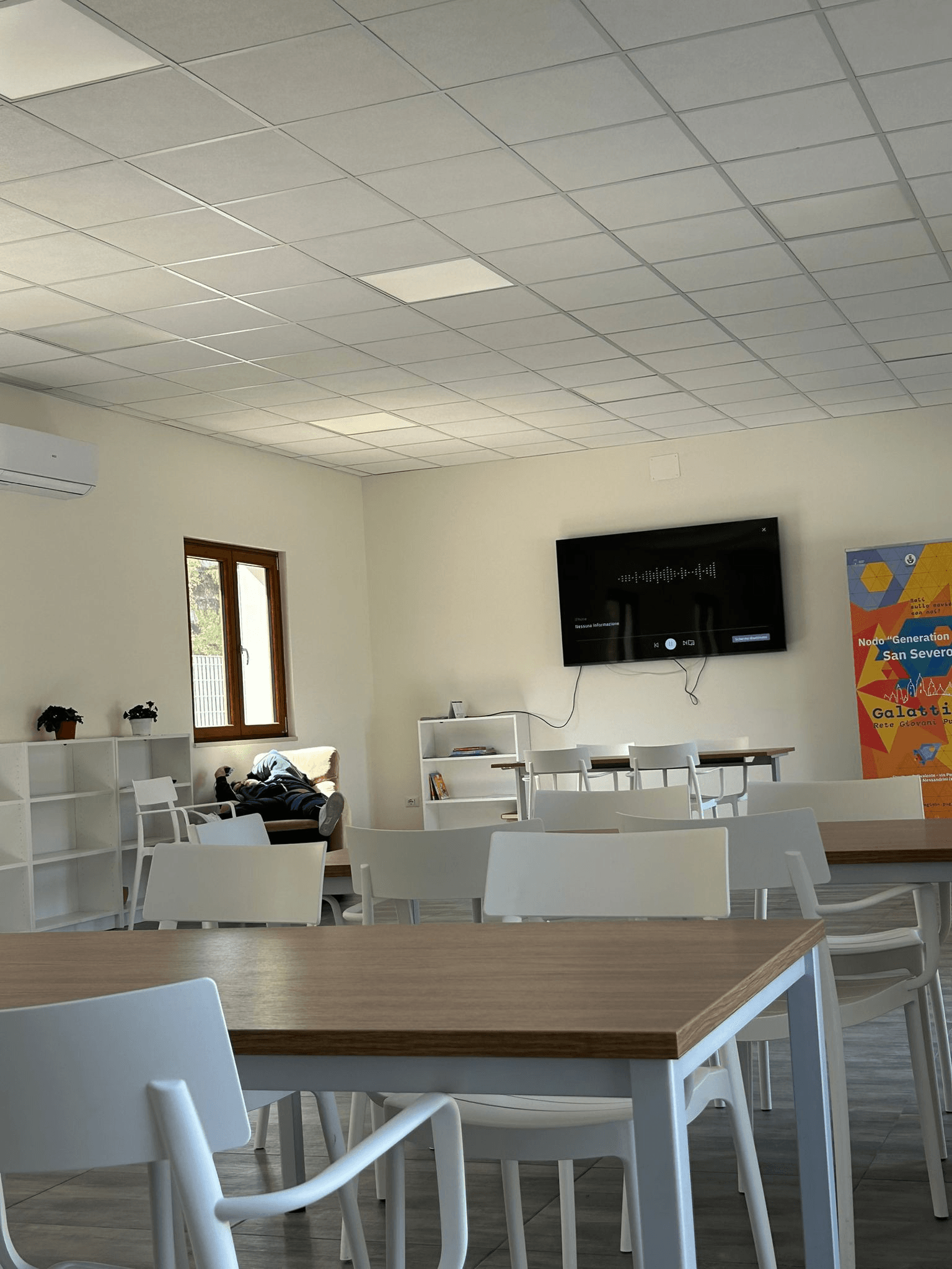 Ampia sala con mura e soffitta chiare, sedie bianche, rtavoli in legno e finestra