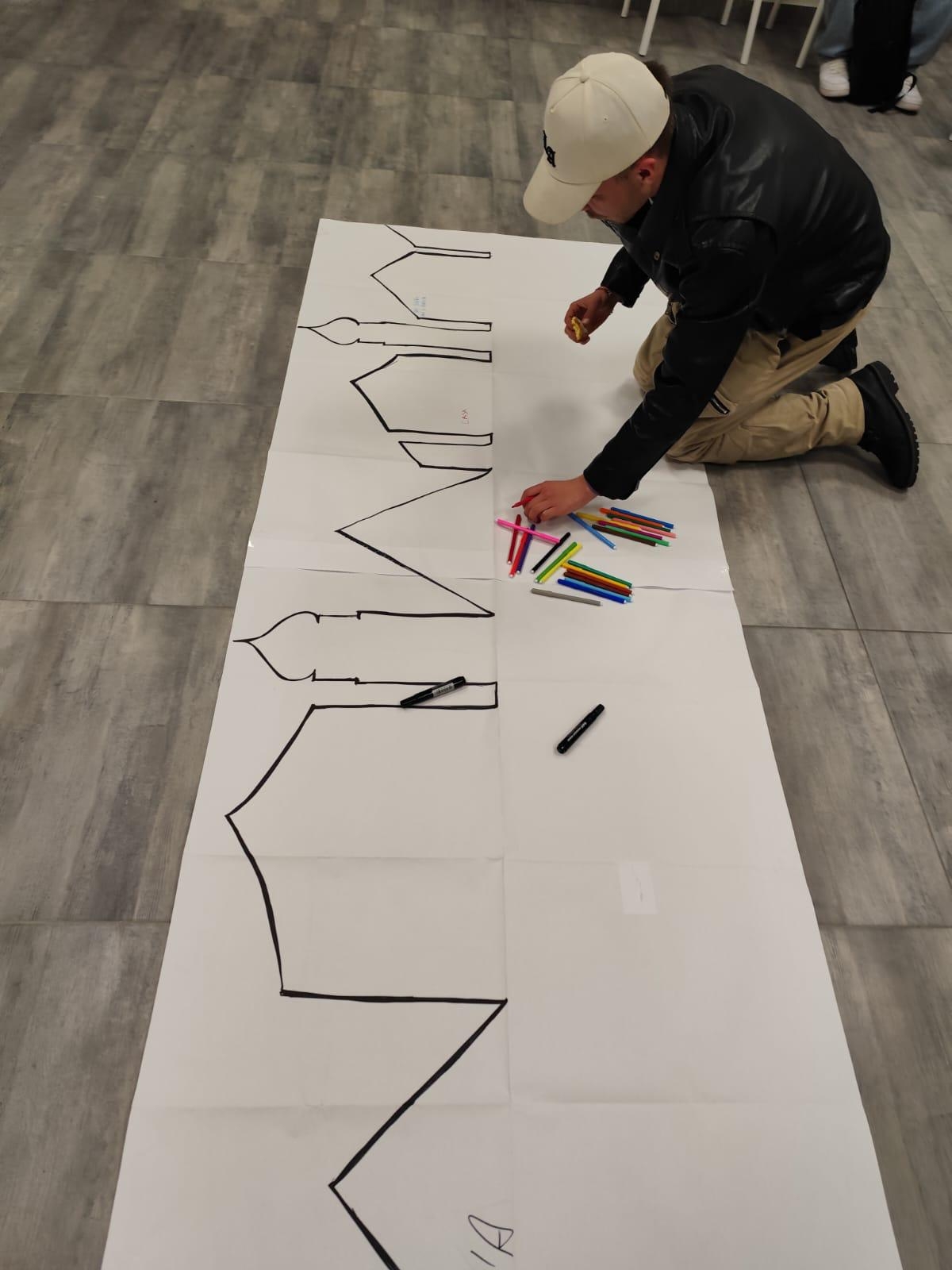 Ragazzo con cappello che scrive su un cartellone bianco predisposto a terra su pavimento scuro