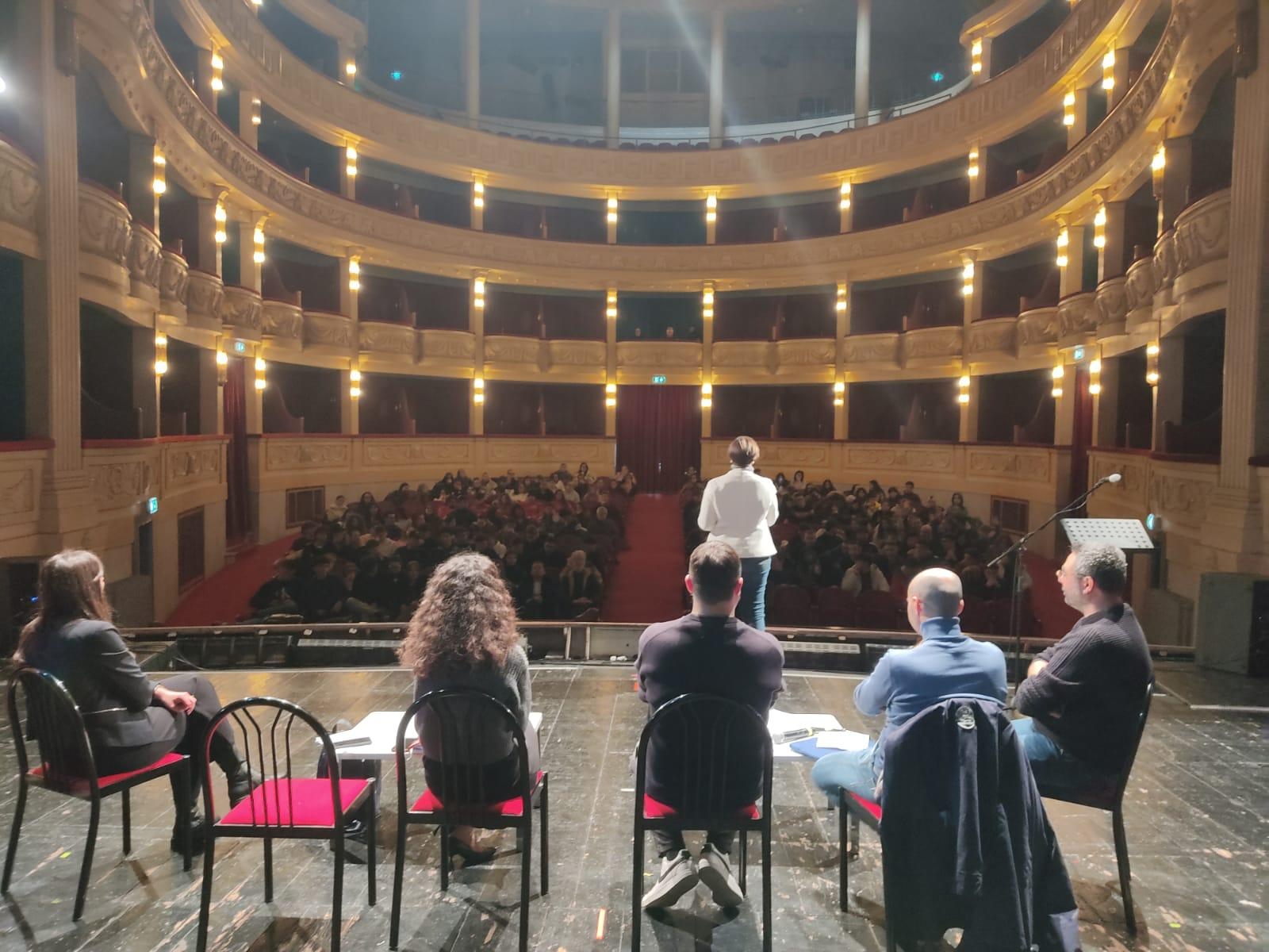 alcune persone sedute su una sedia prediposta su un palco con una donna in piedi di fronte alla platea di un teatro.