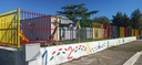 L'entrata dello spazio visto da fuori  Mura bianche con recinzione in ferro colorata e struttura bianca sullo sfondo