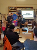 gruppo di giovani partecipanti al workshop di social media manager Giovani seduti intorno ad un tavolo ascoltano il formatore che spiega il workshop di social media manager che si legge in uno schermo proiettato sul muro