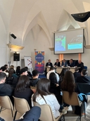 Un gruppo di giovani seduti su delle sedie guarda verso un tavolo centrale e una lavagna digitale sul cui schermo appare locandina dell'evento e la scritta: "Nodo di Galatina: accanto ai giovani per progettare il futuro". Al tavolo sono seduti quattro uomini,di cui il penultimo sulla sinistra sta parlando ad un microfono