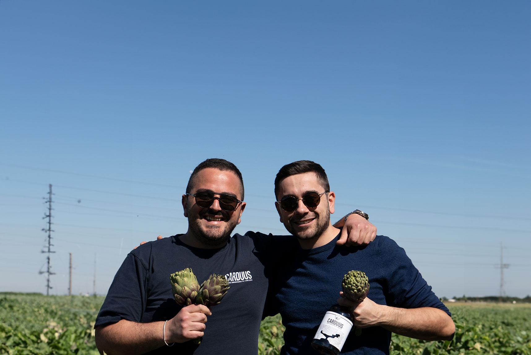 I fondatori di Carduus Due giovani sorridono mentre tengono in mano dei carciofi in un campo all'aperto.
