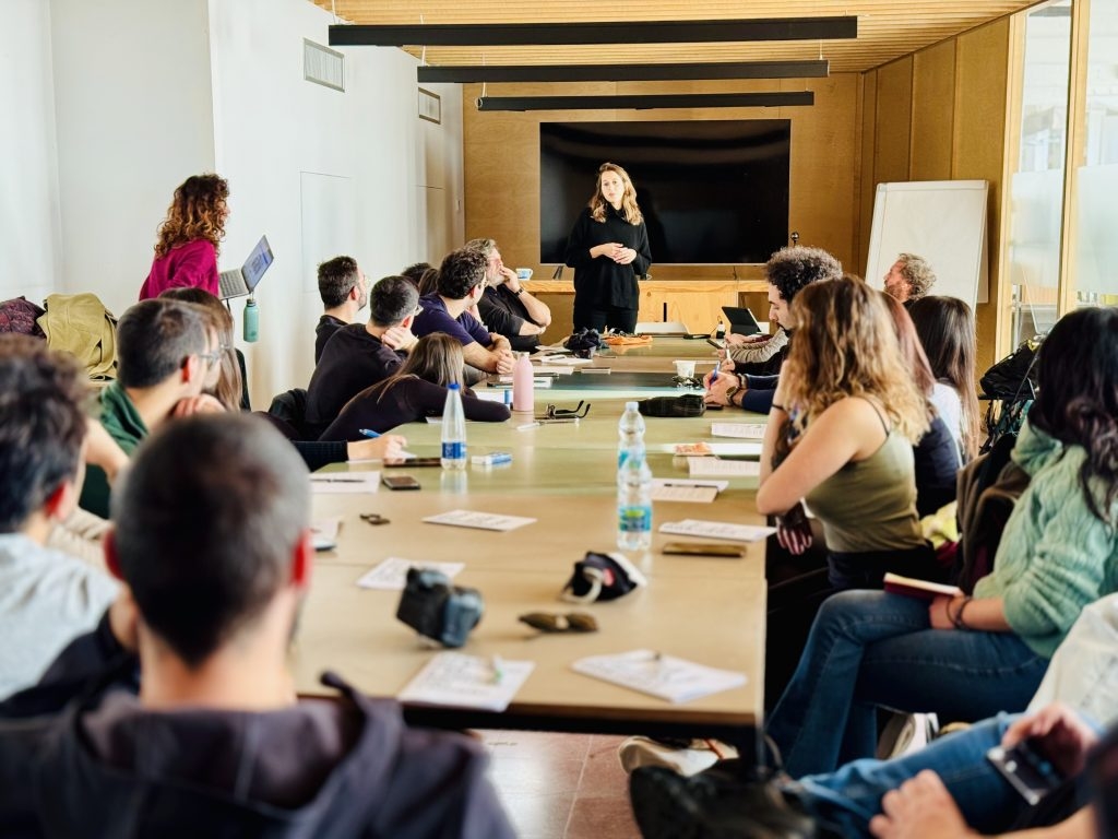 Un gruppo di persone sedute attorno a un lungo tavolo ascolta una relatrice in piedi davanti a uno schermo nero, in una sala con pareti in legno e luce naturale
