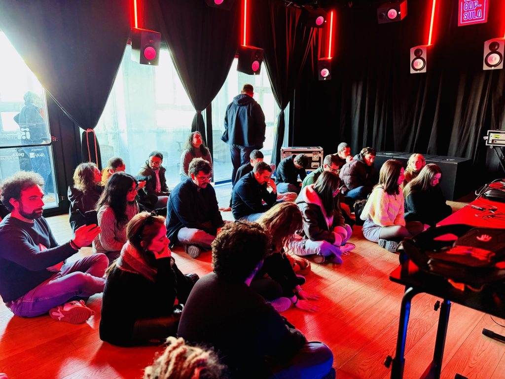 Un gruppo di persone sedute a terra in una sala con luci rosse e nere, circondate da casse audio e tende scure, durante un’esperienza immersiva.
