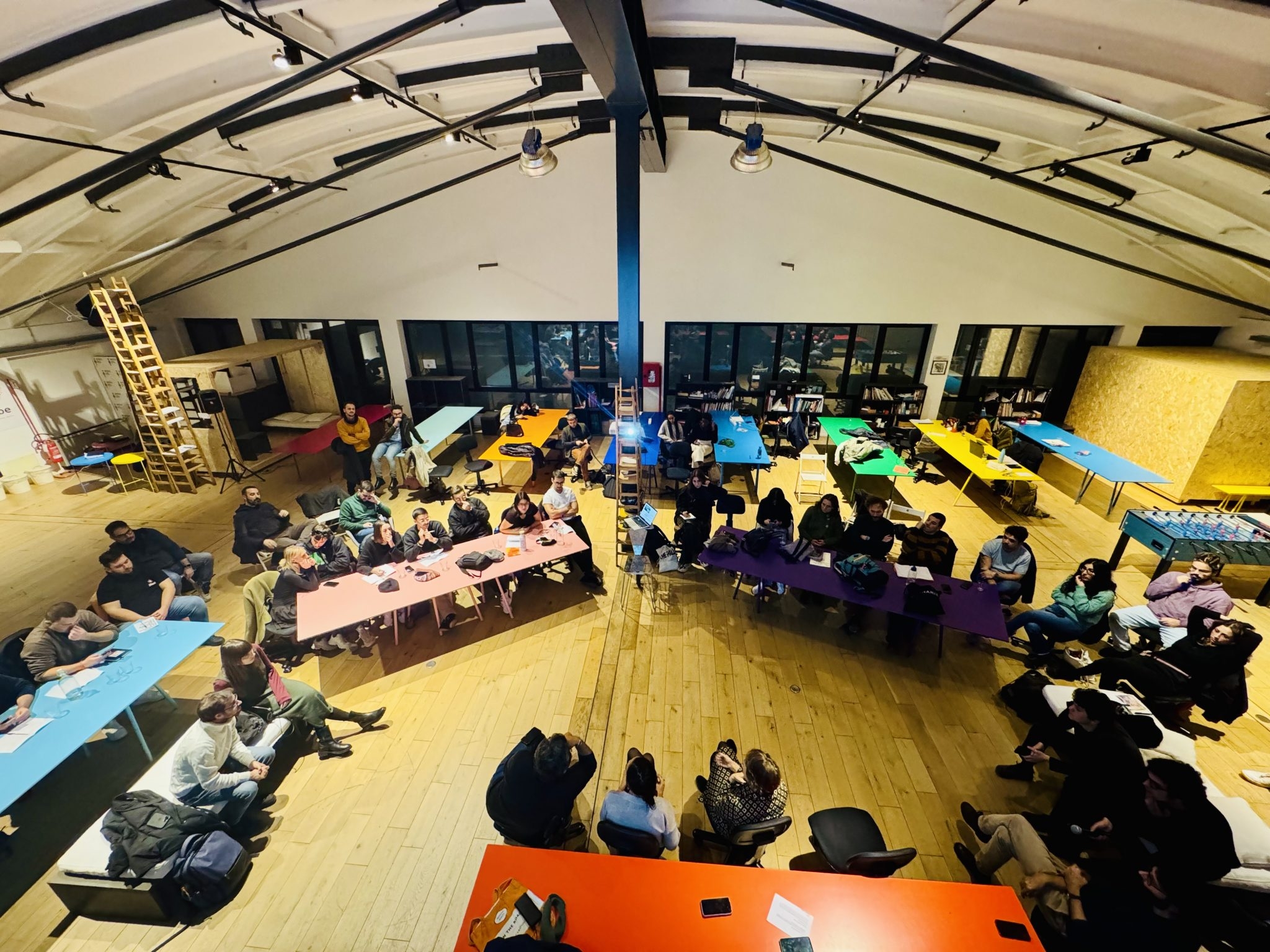 eduta dall’alto di un workshop in uno spazio culturale con partecipanti seduti attorno a tavoli colorati, impegnati in una discussione