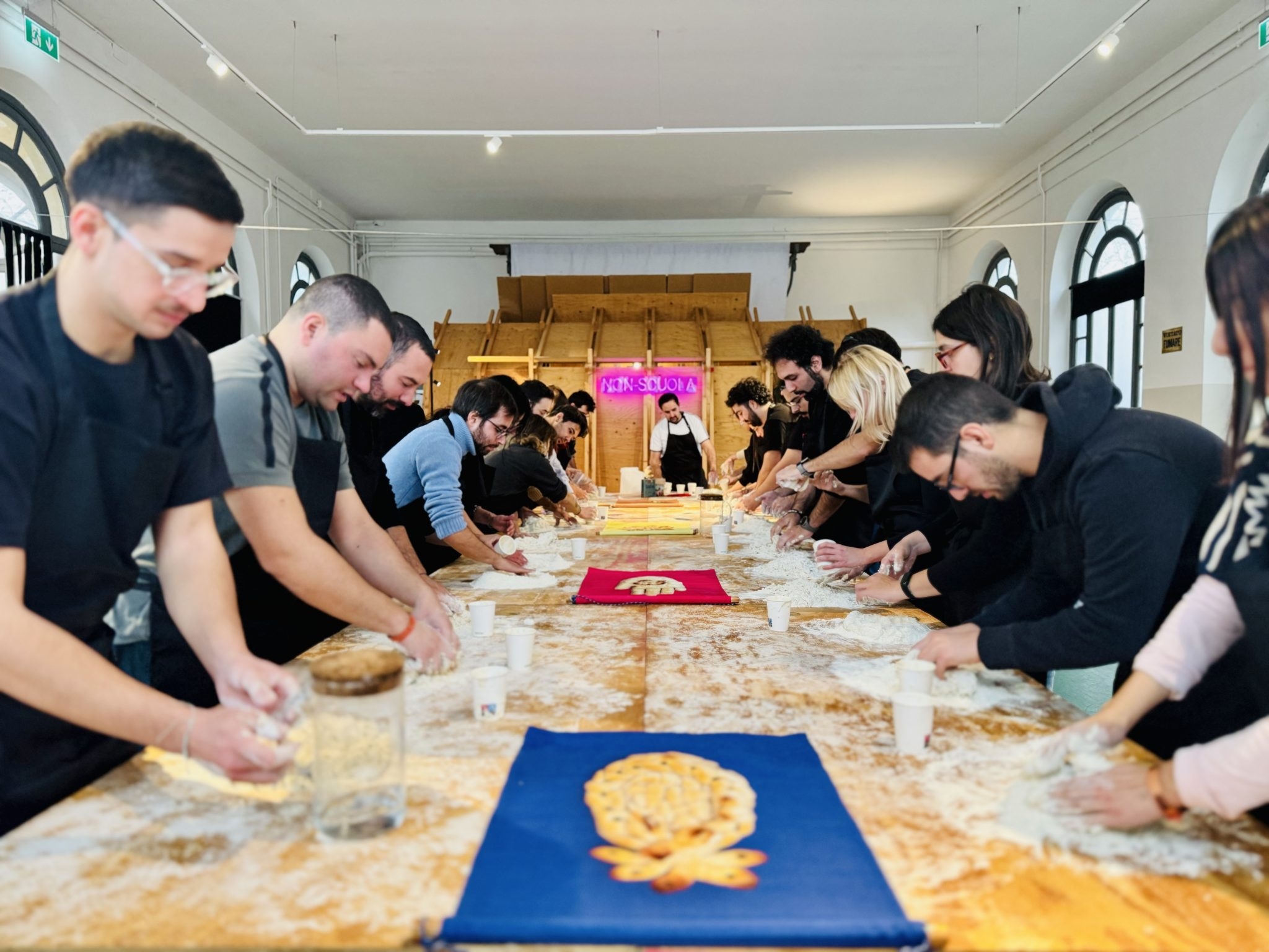 Partecipanti impegnati in un laboratorio di panificazione, lavorando l'impasto su lunghe tavolate in un ambiente luminoso e accogliente