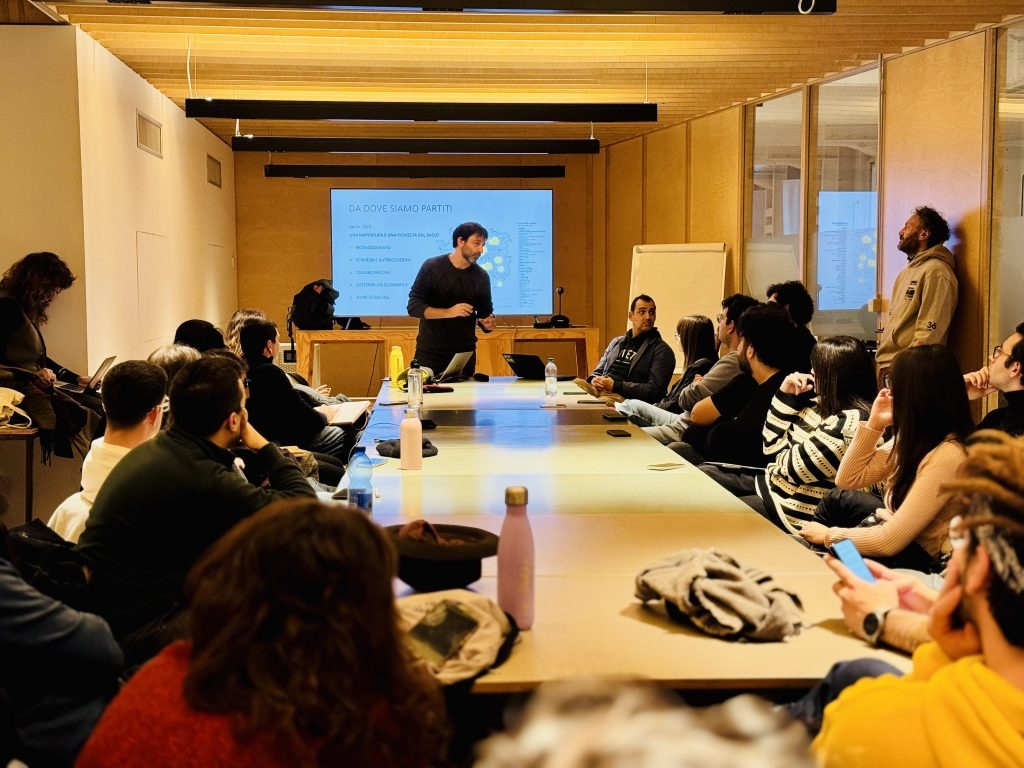 Un uomo in piedi presenta un diagramma su uno schermo a un gruppo di partecipanti seduti attorno a un tavolo, in una sala con pareti in legno e luce naturale