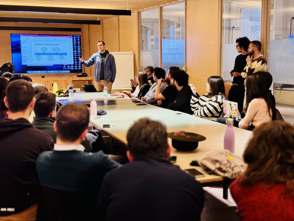 Un uomo in piedi presenta un diagramma su uno schermo a un gruppo di partecipanti seduti attorno a un tavolo, in una sala con pareti in legno e luce naturale