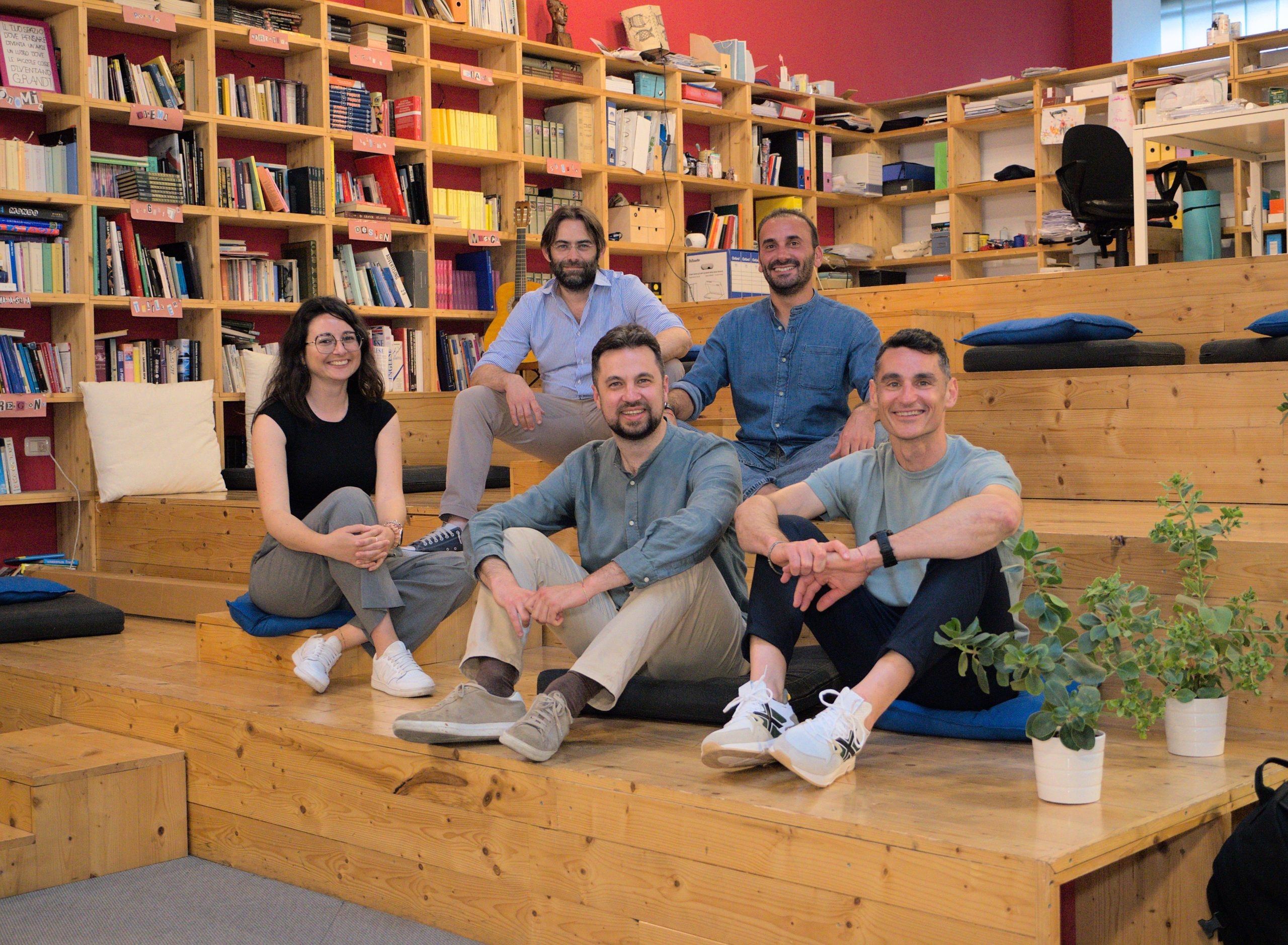 Cinque membri del team Qsensato seduti su gradinate di legno in uno spazio di co-working con scaffali pieni di libri sullo sfondo.