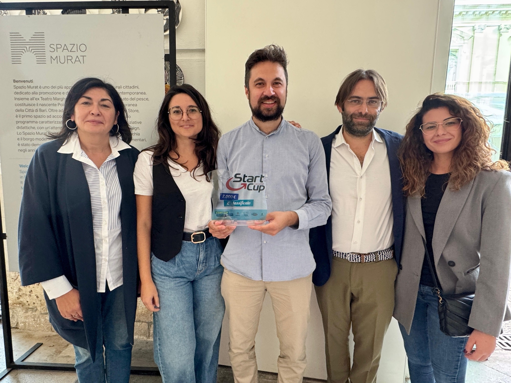 Il team vincitore di Start Cup Puglia 2024 posa con il premio presso lo Spazio Murat di Bari. Cinque persone, tra cui dirigenti e rappresentanti universitari, sorridono mentre tengono il trofeo della competizione.