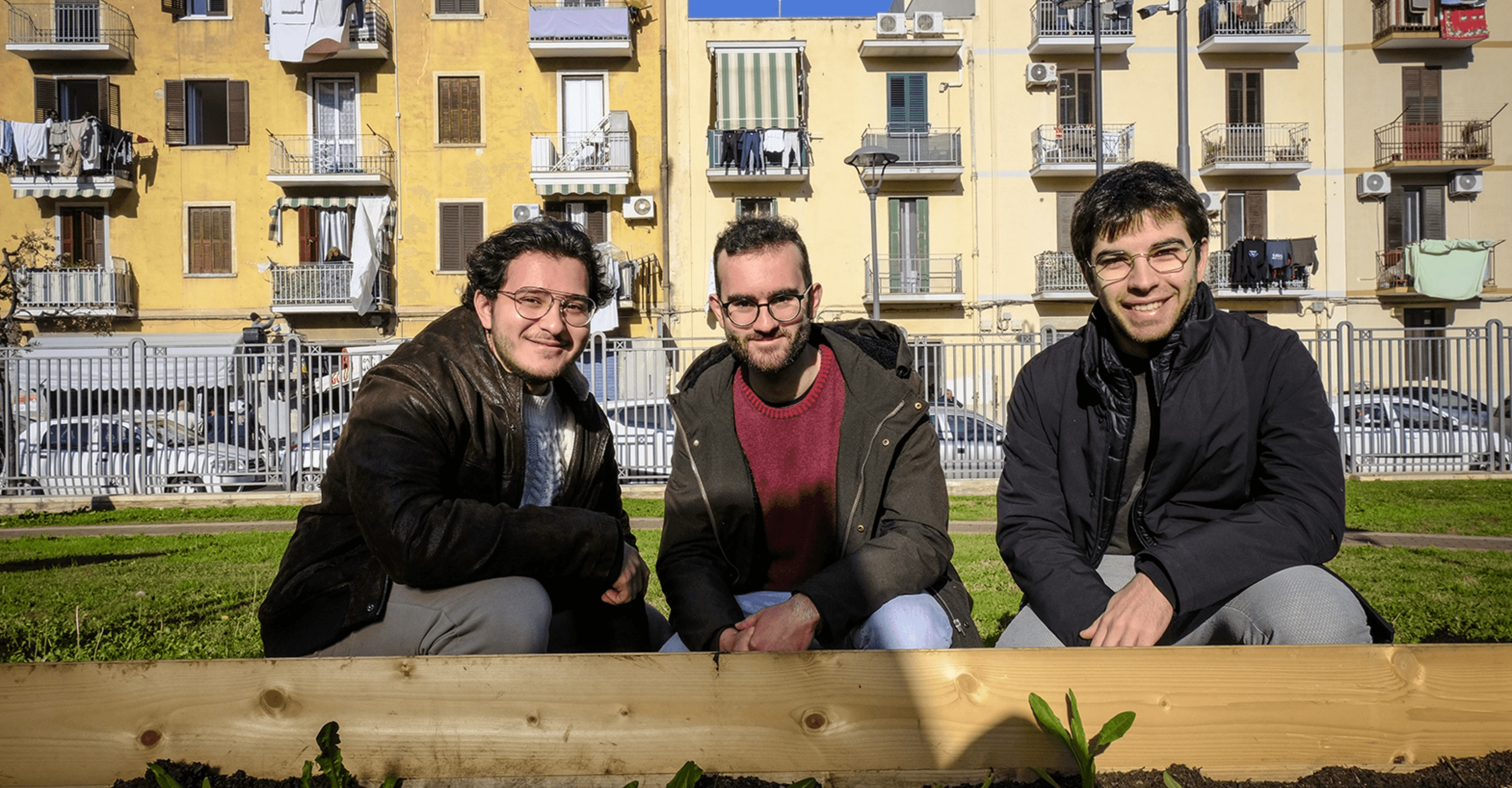 Fridays for Future Bari Libertà – Impegno per il verde urbano Tre giovani attivisti del movimento Fridays for Future Bari sono ritratti accanto a una fioriera in un'area verde del quartiere Libertà. L'iniziativa mira alla riqualificazione urbana attraverso la creazione di spazi verdi condivisi, promuovendo la sostenibilità ambientale e il coinvolgimento della comunità locale. Sullo sfondo, edifici residenziali e automobili che evidenziano il contesto urbano dell'azione.