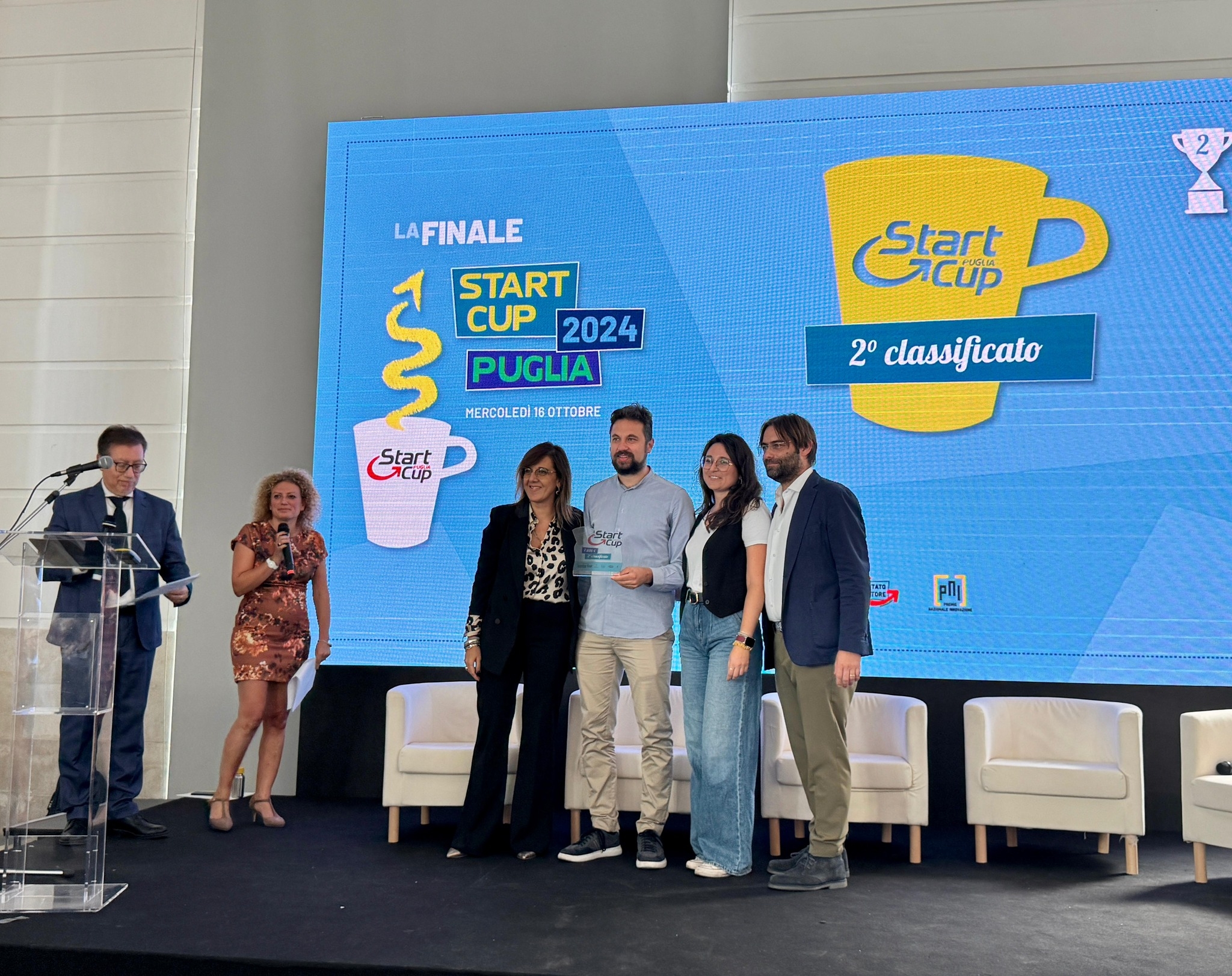 Il team di Qsensato riceve il premio per il secondo posto alla finale di Start Cup Puglia 2024. Sul palco, accanto ai presentatori, il gruppo posa con il trofeo davanti a uno schermo che riporta il logo dell'evento e la dicitura