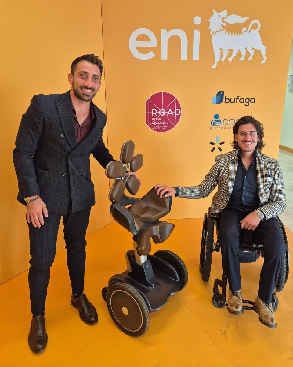 L’immagine mostra Emanuele Battista e Alessio Sansò davanti a una parete arancione con il logo Eni e altri partner tecnologici. Al centro, tra i due, si trova un innovativo dispositivo di mobilità con un design futuristico. Emanuele Battista, in piedi, tiene una mano sul dispositivo, mentre Alessio Sansò, seduto su una sedia a rotelle, appoggia la sua mano sullo stesso. Entrambi sorridono verso la fotocamera.