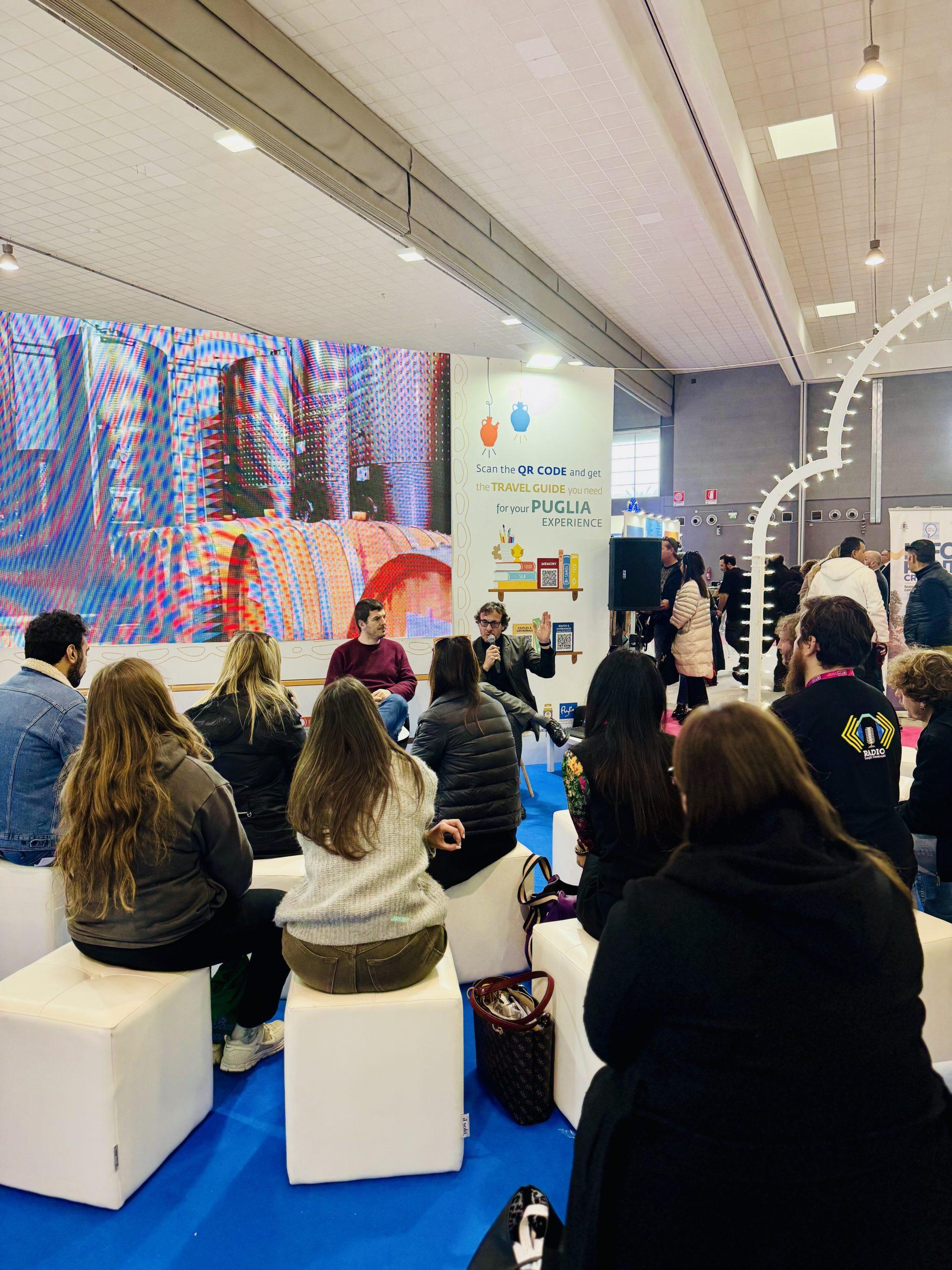 Persone sedute su pouf bianchi assistono a una conferenza durante l'evento BTM 2025, con due relatori che parlano in un'area dedicata alla promozione della Puglia. Sullo sfondo, un grande schermo con immagini colorate e un pannello informativo con QR code per la guida di viaggio della 'Puglia Experience